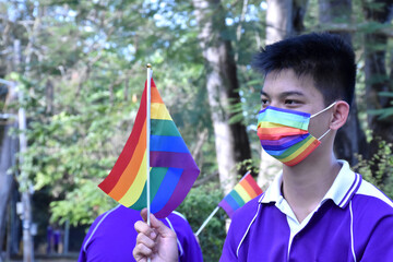 Portrait of young asian boy wears rainbow mask and holding rainbow flag, LGBT symbol, in the park, soft and selective focus, concept for LGBT community celebration and respecting gender diversity.