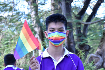 Portrait of young asian boy wears rainbow mask and holding rainbow flag, LGBT symbol, in the park, soft and selective focus, concept for LGBT community celebration and respecting gender diversity.