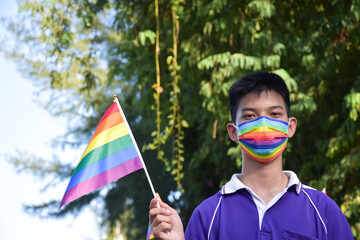Portrait of young asian boy wears rainbow mask and holding rainbow flag, LGBT symbol, in the park, soft and selective focus, concept for LGBT community celebration and respecting gender diversity.
