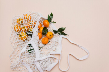 Top view of a white grocery eco-mesh cotton bag with fresh orange tangerines on a beige pastel...