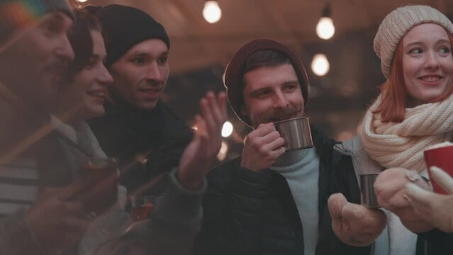 Party vibes. Happy smiling friends with cups of mulled wine having fun, spending time together at winter fair at evening time. Winter holidays