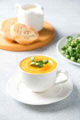 Creamy butternut squash fall soup in elegant white tea cup.Cup of pumpkin cream soup garnished with fresh sunflower microgreen. Front view, selective focus. Vertical image