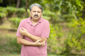 Indian senior man with arm pain standing outdoor at park. Asian Old man hand holding his elbow...