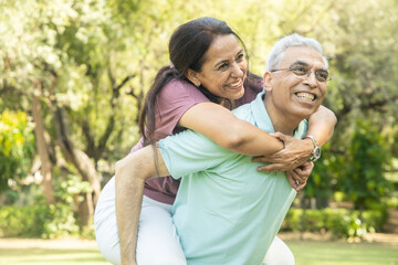 Happy indian senior couple enjoying life having fun at summer park, Aging people, Husband giving piggyback ride to  wife.  Fitness healthcare, Relationship bonding.  - Powered by Adobe