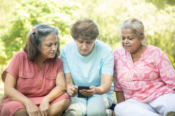 Happy Indian senior women using mobile phone and spend time together outdoor at summer park.