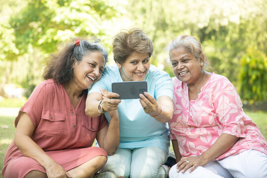 Happy Indian senior women using smart phone and taking pictures of themselves together outdoor at summer park. Elderly people clicking selife .