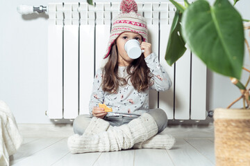 Cute little girl in knitted hat, pajamas, socks having lunch, drink hot tea or cacao and eat sweet cookies near radiator