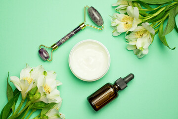 Face roller with cosmetic serum and face cream on a green background with flowers. Facial massage kit for lifting massage.