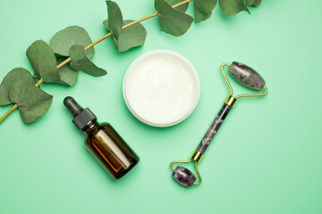 Face roller with cosmetic serum and face cream on a green background with a twig. Facial massage kit for lifting massage.