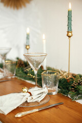 Christmas table setting. Linen napkin with bell on plate, vintage cutlery, glasses, fir branches with golden lights and pine cones on table. Holiday arrangement of table