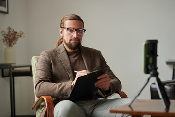 Serious psychologist sitting on armchair in front of mobile phone in tripod and making notes during online consultation
