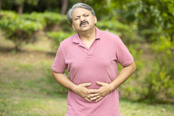 Indian senior man having stomachache or diarrhea outdoor at park, Asian Old male suffering from...