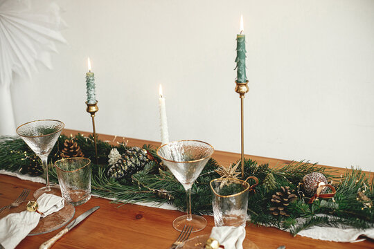 Christmas table setting. Linen napkin with bell on plate, vintage cutlery, glasses, fir branches with golden lights and pine cones on table. Holiday arrangement of table