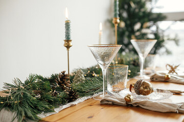 Christmas table setting. Linen napkin with bell, baubles on plate, vintage cutlery, glasses, fir branches with golden lights and pine cones on table. Holiday arrangement of table