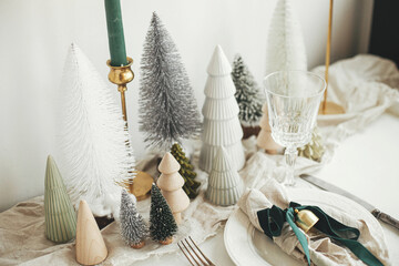Christmas table setting. Stylish napkin with ribbon and bell on plate, vintage cutlery, modern festive christmas trees and houses on white rustic table. Holiday arrangement of table