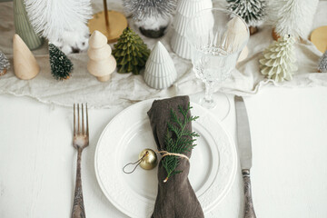 Christmas table setting. Stylish napkin with bell and fir on plate, vintage cutlery, glasses, festive little christmas trees on white rustic table. Holiday arrangement of table, top view