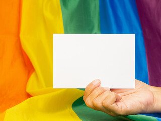 Hand holding a blank white paper against the rainbow flag or LGBT flag background