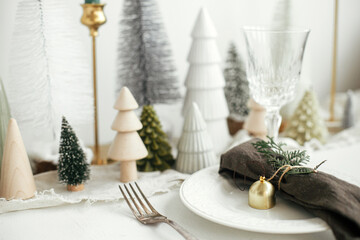 Christmas table setting. Stylish napkin with bell and fir on plate, vintage cutlery, glasses, festive little christmas trees on white rustic table. Holiday arrangement of table