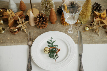 Christmas luxury table setting. Cedar branch with bell on plate, vintage cutlery, glasses, golden little christmas trees and ornaments on table. Holiday arrangement of table, top view