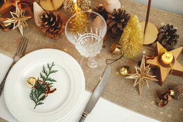 Christmas luxury table setting. Cedar branch with bell on plate, vintage cutlery, glasses, golden little christmas trees and ornaments on table. Holiday arrangement of table