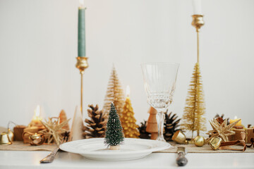 Stylish Christmas table setting. Little christmas tree on plate, vintage cutlery, wineglass, modern golden christmas trees and candles on table. Holiday brunch, new year celebration