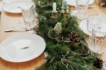 Stylish Christmas table setting. Rosemary branch on plate, vintage cutlery, wine glass, candles, fir branches with golden lights, pine cones on table. Holiday dinner, atmospheric table set