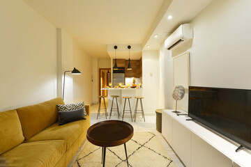 Living room of a vacation rental apartment with white wooden furniture, a three-seater sofa in yellow fabric, a circular coffee table and a kitchen table with high stools