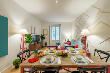 Rectangular wooden dining table with tableware set with fresh fruit, sweets and pastries and glasses of orange juice in a decorated living room