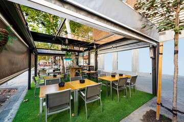 Terrace of a bar with a floor covered with artificial grass, wooden tables with gray armchairs and...