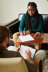 African American patient having art therapy at consultation with psychologist, she sitting at table...