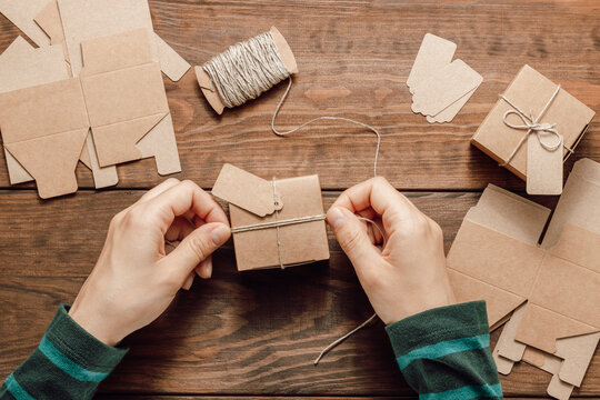 Hands Wrap Paper Gift Box With Twine Ribbon On Wooden Background. Woman Makes Gift Box With Mockup Paper Label. DIY Gift Box. Holidays Preparation, Handmade Gift Box, Top View, Flat Lay