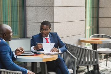 Two dark-skinned African American businessmen in suits and glasses with briefcases sit at a table in an outdoor city cafe.