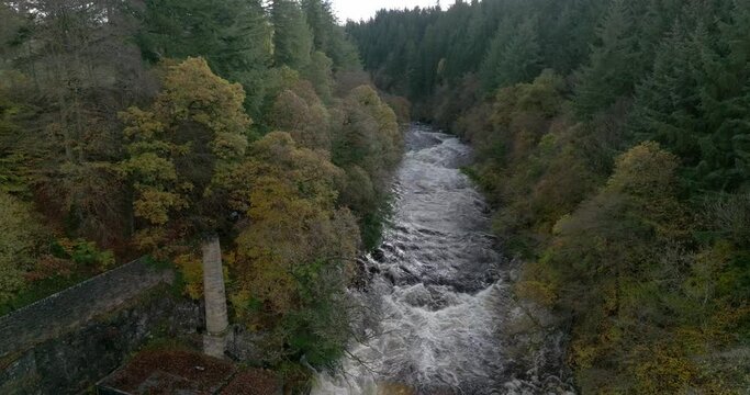 Drone footage reverses down a fast flowing river and waterfall surrounded by old buildings and a forest of broadleaf and coniferous trees. Falls of Clyde, New Lanark, Scotland.