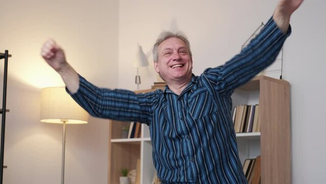 Weekend joy. Happy aged man. Home rest. Cheerful funny elder male in pajamas celebrating hands up in light room interior slow motion.