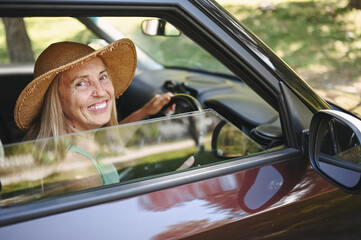 Naklejka premium Happy senior woman driver in straw hat driving sitting in new car, smiling looking at camera enjoying journey. Driving courses and life insurance. Retired people activity and road trip concept 