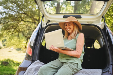 Naklejka premium Senior happy woman working with laptop from the car trunk or boot in camper sunny summer park - digital nomad freedom lifestyle concept, travel freelancer and vacations of retired old age people.