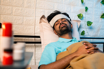 young indian sick man suffering from mild fever while sleeping at night by placing cold cloth on forehead - concept of healthcare, covid symptoms and illness.
