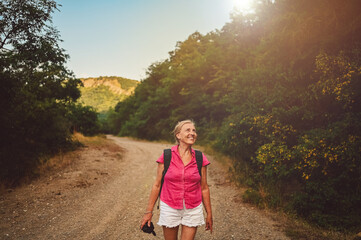 Excited happy senior woman backpacker tourist walking in summer forest road outdoors at sunset time. Old slim lady traveling with photo camera. Active retirement vacation concept. Warm filter