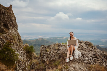 Young woman freelancer traveler working online anywhere outdoors using laptop enjoying mountain peak view. Happy female downshifter holding computer in unusual amazing view workplace at summer