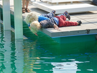 Zwei hell blonde Kinder liegen auf einem Holzsteg und schauen hinab ins klare grüne blaue Wasser...