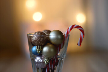 Glass with colorful Christmas baubles and candy cane. Lit candles and Christmas lights in the background. Selective focus.