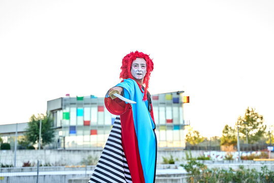 Stilt Walker In Colorful Costume Holding A Sword Looking At The Camera In The Open Air With A Colorful Building In The Background.