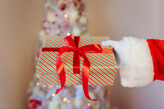 Santa Claus's hand in a white glove holds a gift box on the background of a decorated Christmas tree - Powered by Adobe