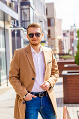 A young man dressed in a beige coat is walking down the street. A young bearded guy with a modern hairstyle and sunglasses on an urban background