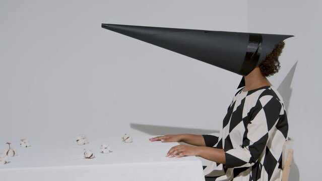 Horizontal side view studio shot footage of unrecognizable young woman wearing black and white outfit with conus-shaped mask on face sitting at table with cotton flowers on it