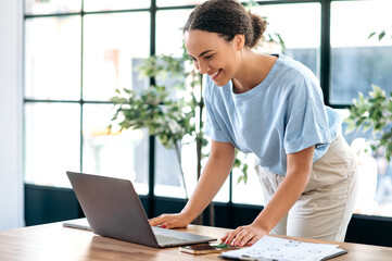 Positive concentrated busy mixed race female employee, successful business lady, working with laptop, typing text, answering email, while standing in modern office near work desk, smiling