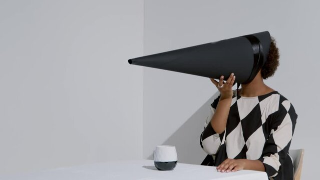 Horizontal conceptual shot footage of unrecognizable African American woman wearing black and white outfit with conus-shaped mask on face sitting at table