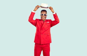Joyful African American man in stylish outfit isolated on light blue background. Happy cheerful good looking young guy in red suit and sunglasses smiles and puts on trendy panama hat. Fashion concept