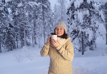 Fototapeta na wymiar happy mature woman 50 years old drink hot tea in winter forest, early evening in park among snow-covered trees, beautiful landscape, smiling happily, concept active lifestyle, enjoying winter nature