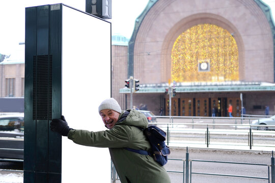funny mature man in winter clothes cheerfully pushes empty blank billboard near Helsinki station square, Helsinki City Transport, winter street, Helsinki, Finland, basis for designer